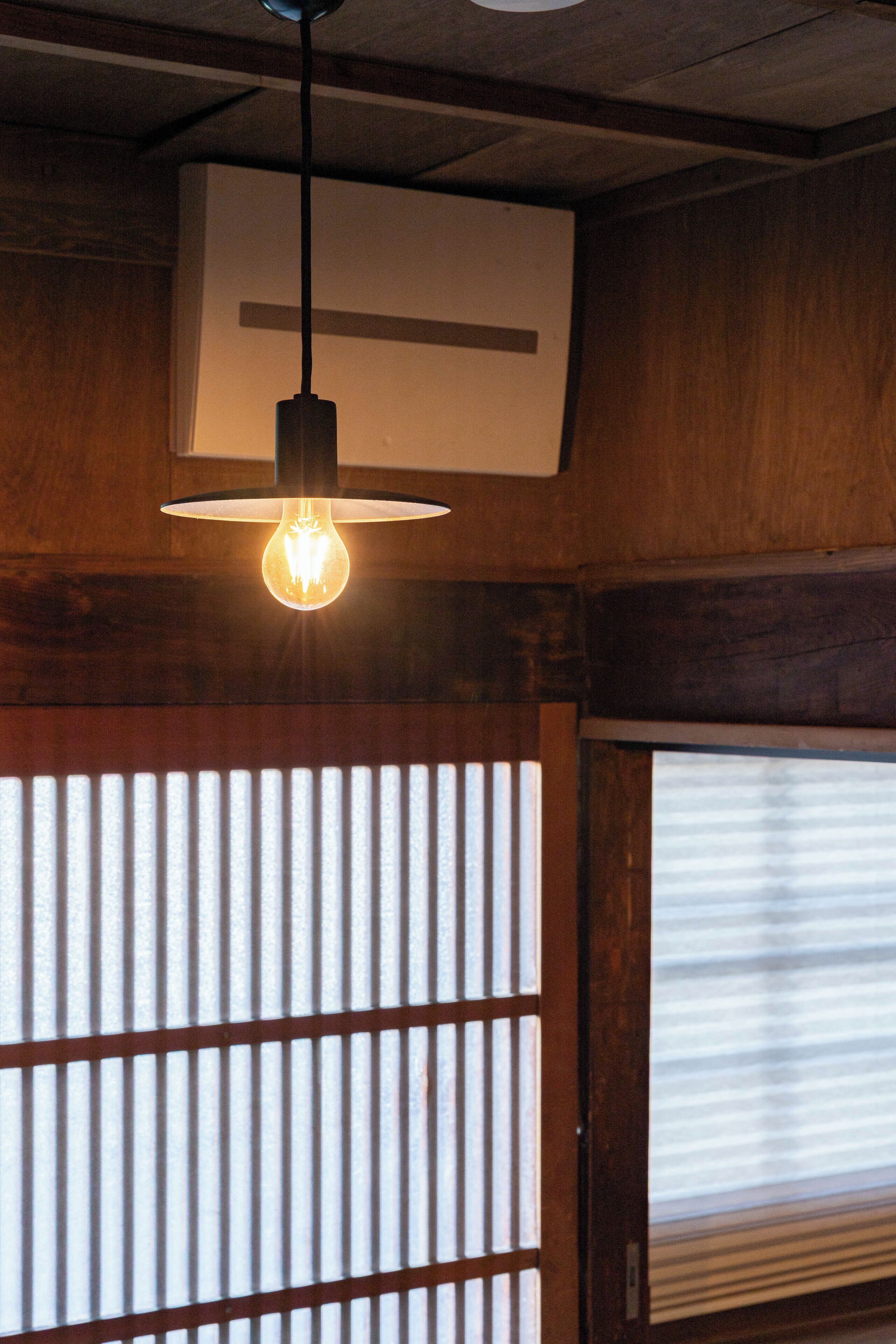 A hanging light fixture with a visible bulb, a wooden wall panel, and a window with blinds.