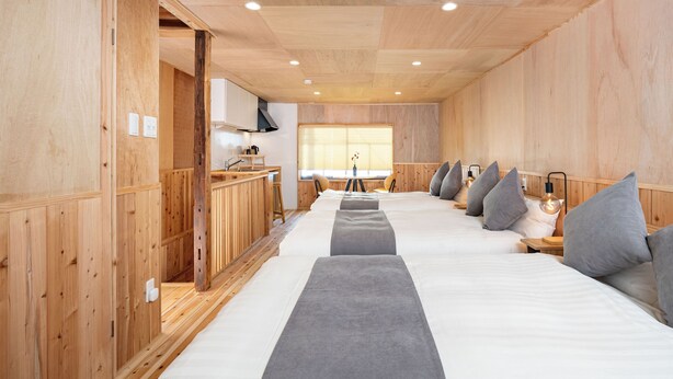 A wooden interior with multiple beds aligned along a central walkway, a kitchenette area, and a window with blinds. - Mameyama15 (Nara)
