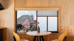 A room with a round wooden table, two chairs, a vase with flowers, and a window with a view of traditional architecture and cherry blossoms. - Mameyama15 (Nara)
