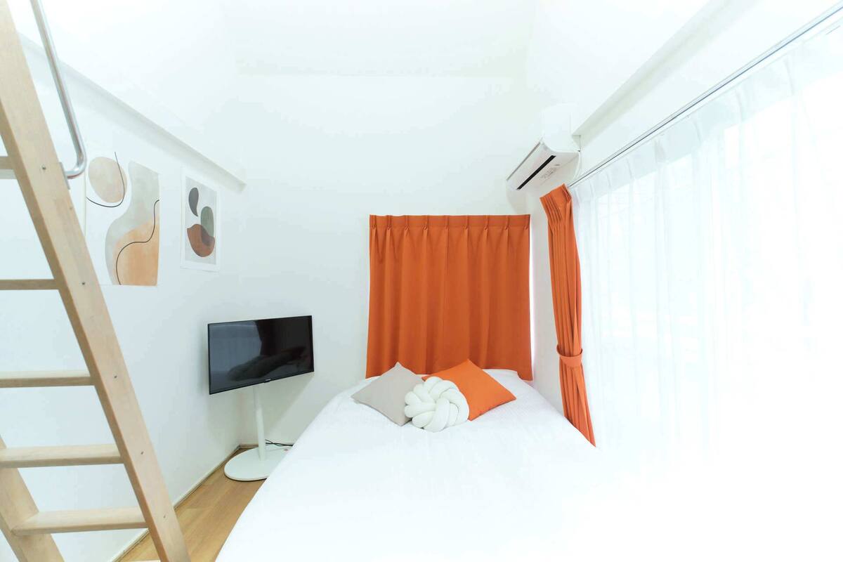A bedroom with a bed, a TV, a ladder, and a window with curtains.