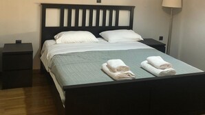 A neatly made bed with white linens and a grey bedspread, two bedside tables, a wall-mounted lamp, and a painting of a person. - Luxurious Apartment With big Balcony Best Location (Athens)