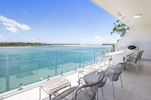A balcony with a view of a beach and ocean, furnished with wicker chairs and a table, and a barbecue grill. - River's Edge At Mariners (Noosa)