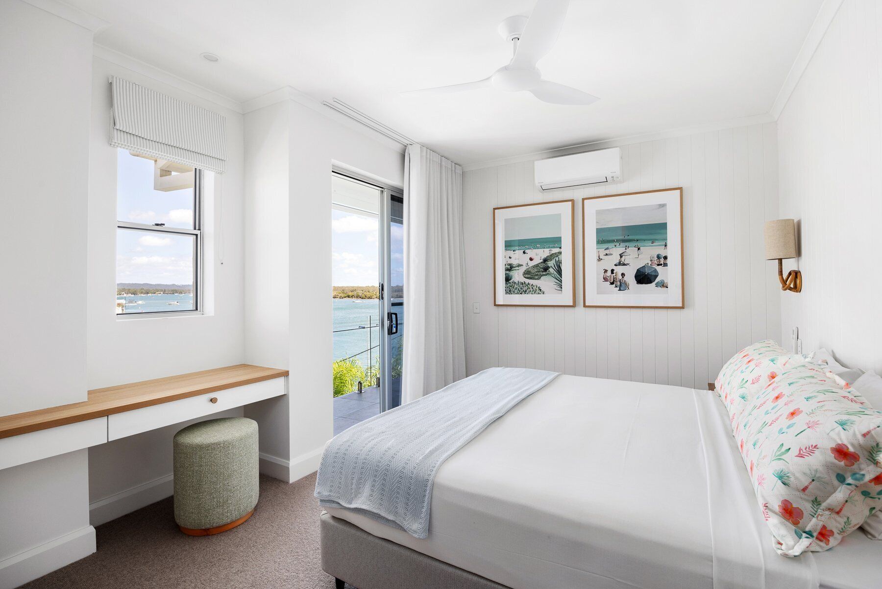 A bedroom with a bed, a desk, a chair, a ceiling fan, and a window with a view of the sea.