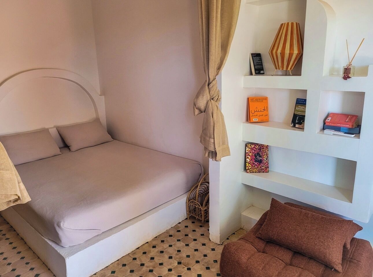 Een slaapkamer met een bed, een kleine bank, een plank met boeken en decoratieve objecten, en een kamer met een patroon op de vloer.