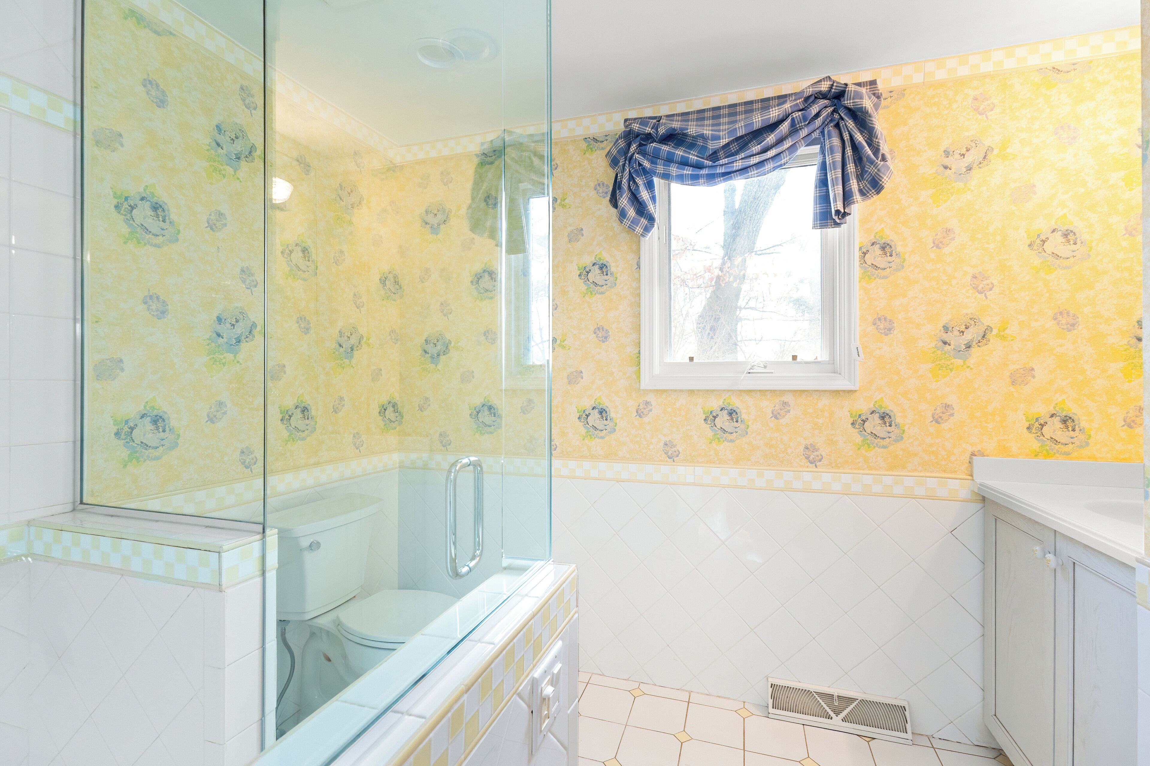 Et badeværelse med toilet, glasvæg til brusekabine og et vindue med ternet gardin.