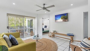 A modern living room with a yellow sofa, a round coffee table, and a ceiling fan. There is a large window with a view of greenery outside.