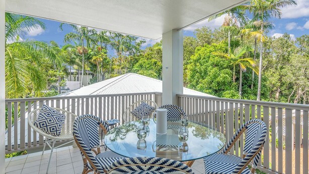 A glass-top table with six chairs, a white railing, and a view of tropical trees.