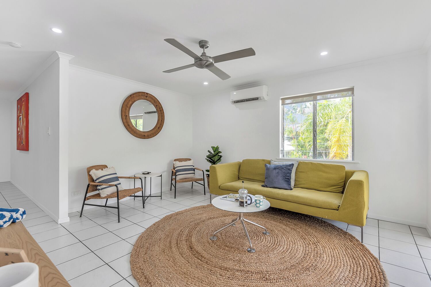 Ein modernes Wohnzimmer mit einem gelben Sofa, zwei Sesseln, einem runden Couchtisch, einem Deckenventilator und einem großen Fenster mit Blick auf Grünflächen.