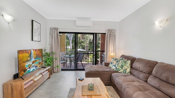 A modern living room with a brown leather sofa, a wooden coffee table, a TV on a wooden stand, and a balcony with a table and chairs.