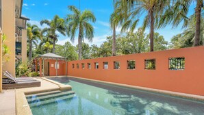 A swimming pool with clear water, surrounded by a low orange wall, palm trees, and a building with multiple windows.