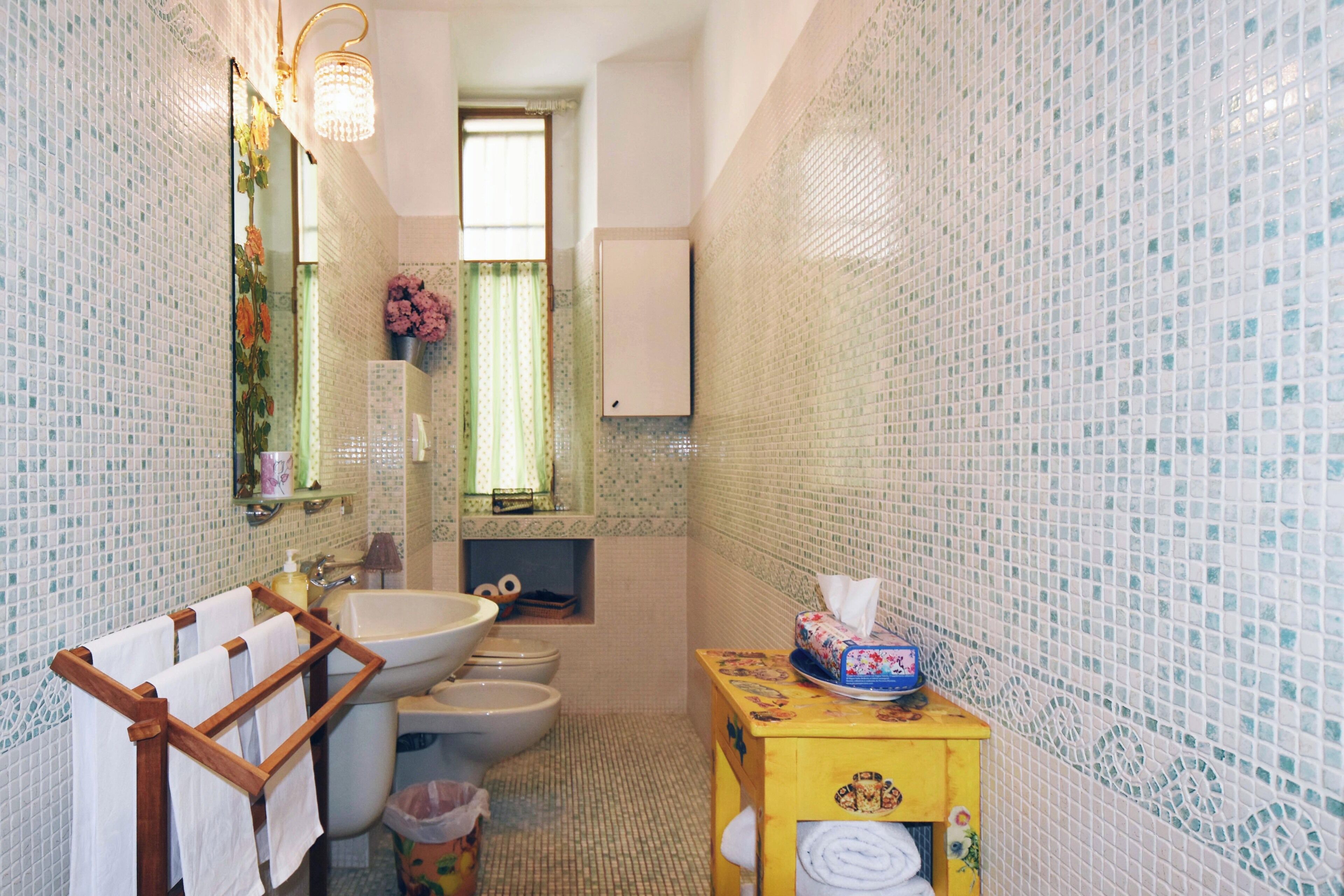 Une salle de bain équipée d’un lavabo, de toilettes et d’un miroir. On y trouve un porte-serviettes avec des serviettes, une poubelle et une boîte décorative sur laquelle est posée une boîte à mouchoirs.