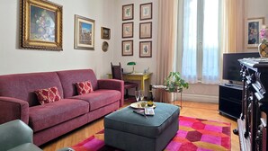 Un soggiorno con un divano rosso, un poggiapiedi grigio e un mobile TV. Alle pareti ci sono quadri incorniciati e una finestra con tende.