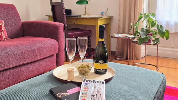 Un soggiorno con un divano rosso, un tavolino con una bottiglia di Prosecco, bicchieri e una ciotola di olive. Sulla tavolino è presente una rivista di Milano.