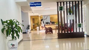 Uma área de recepção com uma placa indicando 'Reception', um recepcionista e um balcão de atendimento. Há plantas em vasos e um vaso grande com um logotipo.