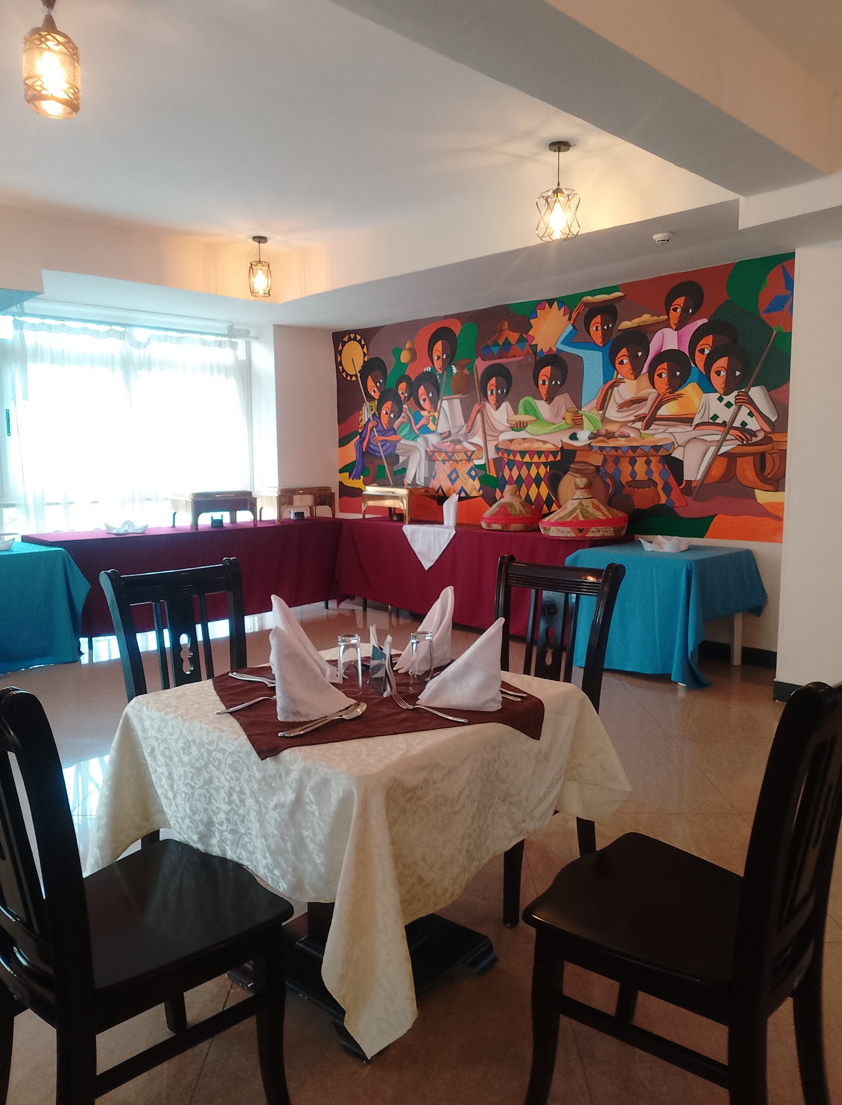 Un comedor con mesas dispuestas para cuatro personas, sillas y un gran mural en la pared.