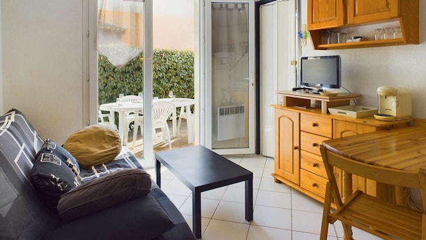 Un espace de vie compact comprenant un canapé, une petite table, un meuble en bois avec un téléviseur et une machine à café, ainsi qu’une porte-fenêtre coulissante donnant sur un espace extérieur avec une table et des chaises.