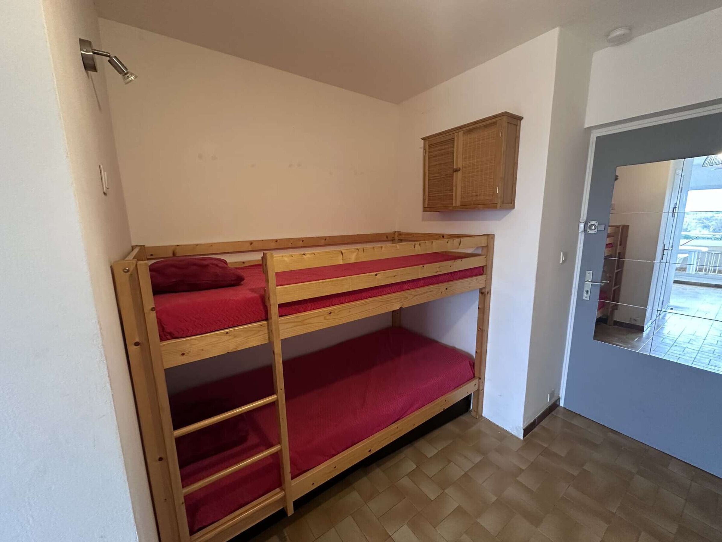Una cama litera con ropa de cama roja, un armario de madera arriba y una puerta que da a un espacio exterior.