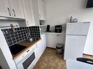 Una cocina compacta con gabinetes blancos, salpicadero de baldosas negras, horno empotrado, cafetera, refrigerador y un bote de basura.