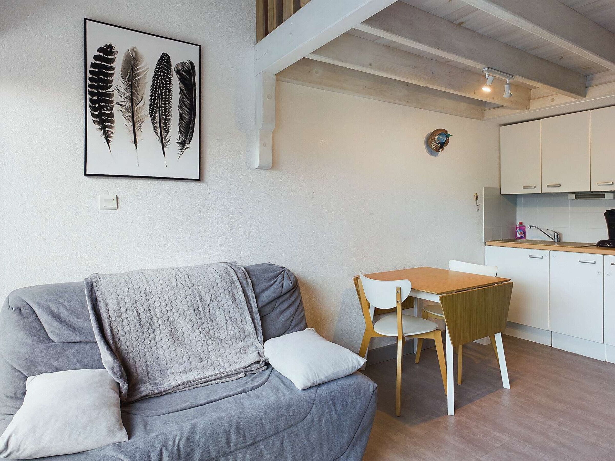 A modern living room with a grey sofa, a wooden dining table, and a kitchen area with white cabinets.