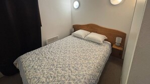 A small, neatly arranged bedroom with a single bed, a bedside table with a lamp, and a wall-mounted light fixture.