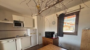 A compact kitchen with a wooden table and chairs, a microwave, a refrigerator, and a window with a view of buildings.