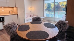 A modern kitchen with a built-in oven, a dining table with four dark placemats, a fluffy chair, and a sofa with a view of trees through a large window. - Perfect studio for 4, ski-in/ski-out with large terrace (Les Deux Alpes)