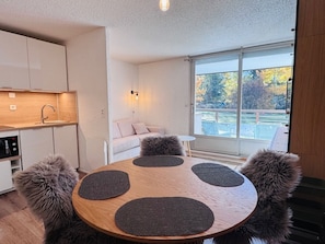 A modern kitchen with a built-in oven, a dining table with four dark placemats, a fluffy chair, and a sofa with a view of trees through a large window.
