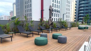 Terraza en la azotea con sillones, otomanas y sombrillas, rodeada de edificios altos.