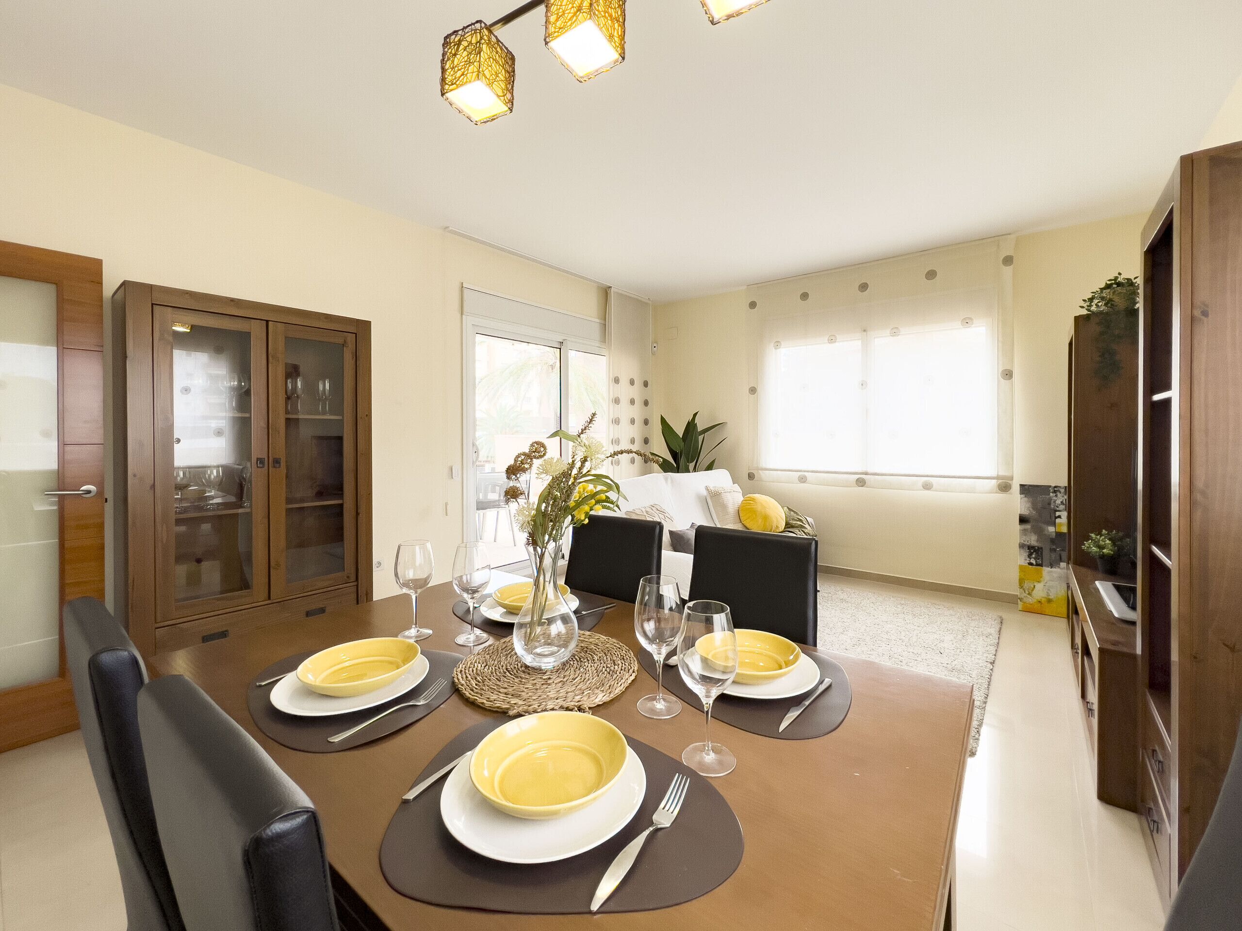 Un coin repas avec une table dressée pour quatre personnes, des chaises, un buffet aux portes vitrées et une fenêtre avec des stores.
