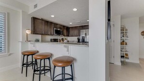 Een moderne keuken met donkere houtkasten, een roestvrijstalen koelkast en een wit aanrecht. Er staan drie krukken met leren zittingen.