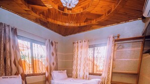 Una habitación con dos camas de madera, un banco de madera, una mesita pequeña de madera y una ventana con cortinas.