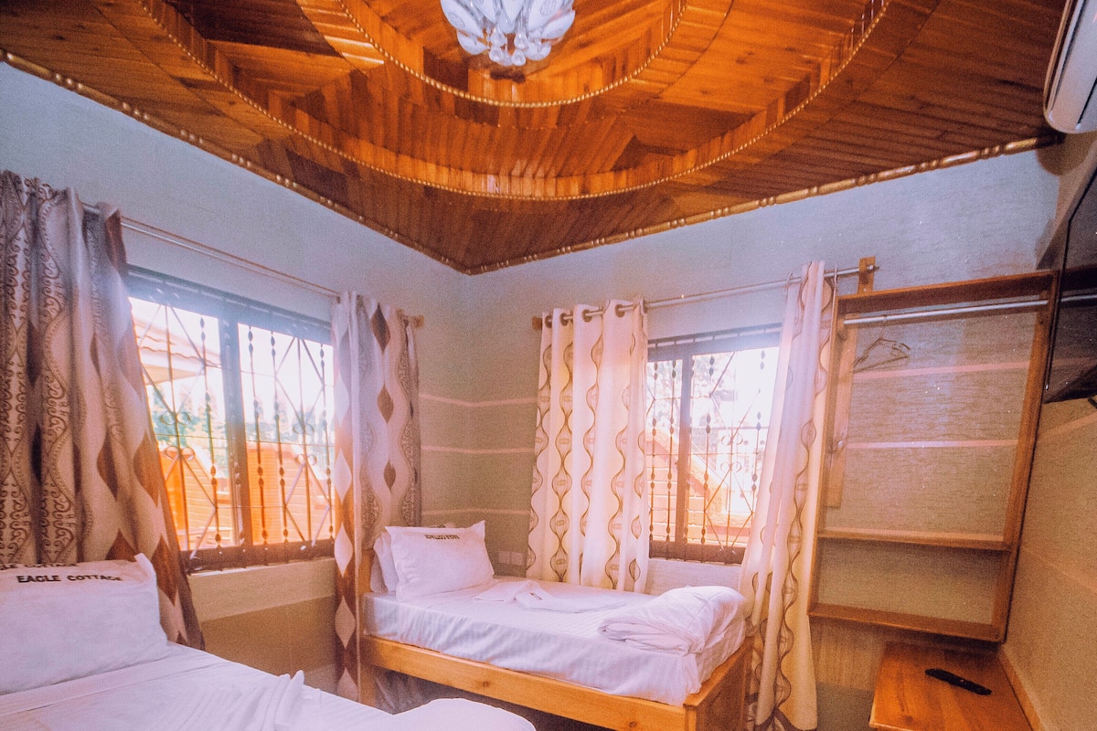 Una habitación con dos camas de madera, un banco de madera, una mesita pequeña de madera y una ventana con cortinas.