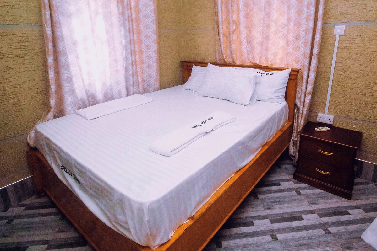 Una cama bien hecha con estructura de madera y sábanas blancas, una mesita de noche de madera con una lámpara, y un suelo con diseño.