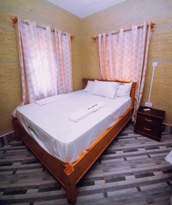 A neatly made bed with a wooden frame and white linens, a small wooden nightstand with a lamp, and a patterned floor.