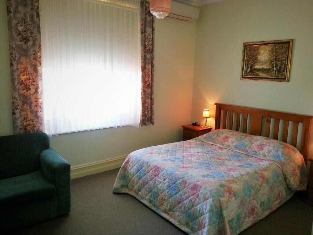 A bedroom with a bed, a chair, a nightstand with a lamp, a painting on the wall, and a window with curtains.