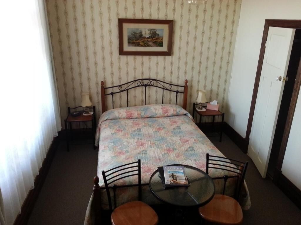 A single bed with a floral bedspread, a bedside table with a lamp, a framed picture on the wall, and a small round table with chairs in the foreground.