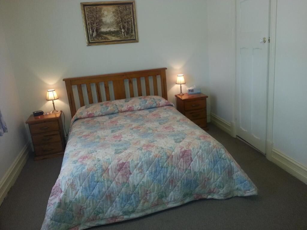Une chambre avec un lit en bois, une literie à motifs floraux, des lampes de chevet, une table de nuit, un tableau encadré au mur et une porte.