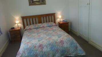 Une chambre avec un lit en bois, une literie à motifs floraux, des lampes de chevet, une table de nuit, un tableau encadré au mur et une porte.
