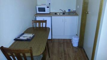 A compact kitchen with a microwave, sink, and cabinets. There is a dining table with chairs and a trash bin.