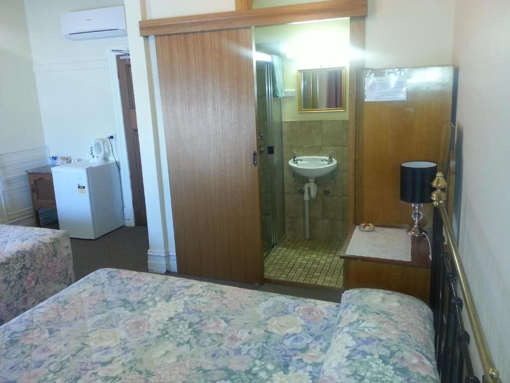 A hotel room with a bed, a small desk with a lamp, a shower area, and a refrigerator.