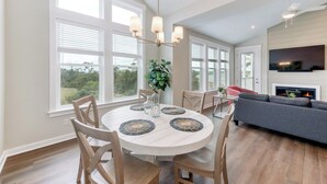A dining area with a round table, chairs, and a chandelier. There is a sofa and a fireplace in the background.