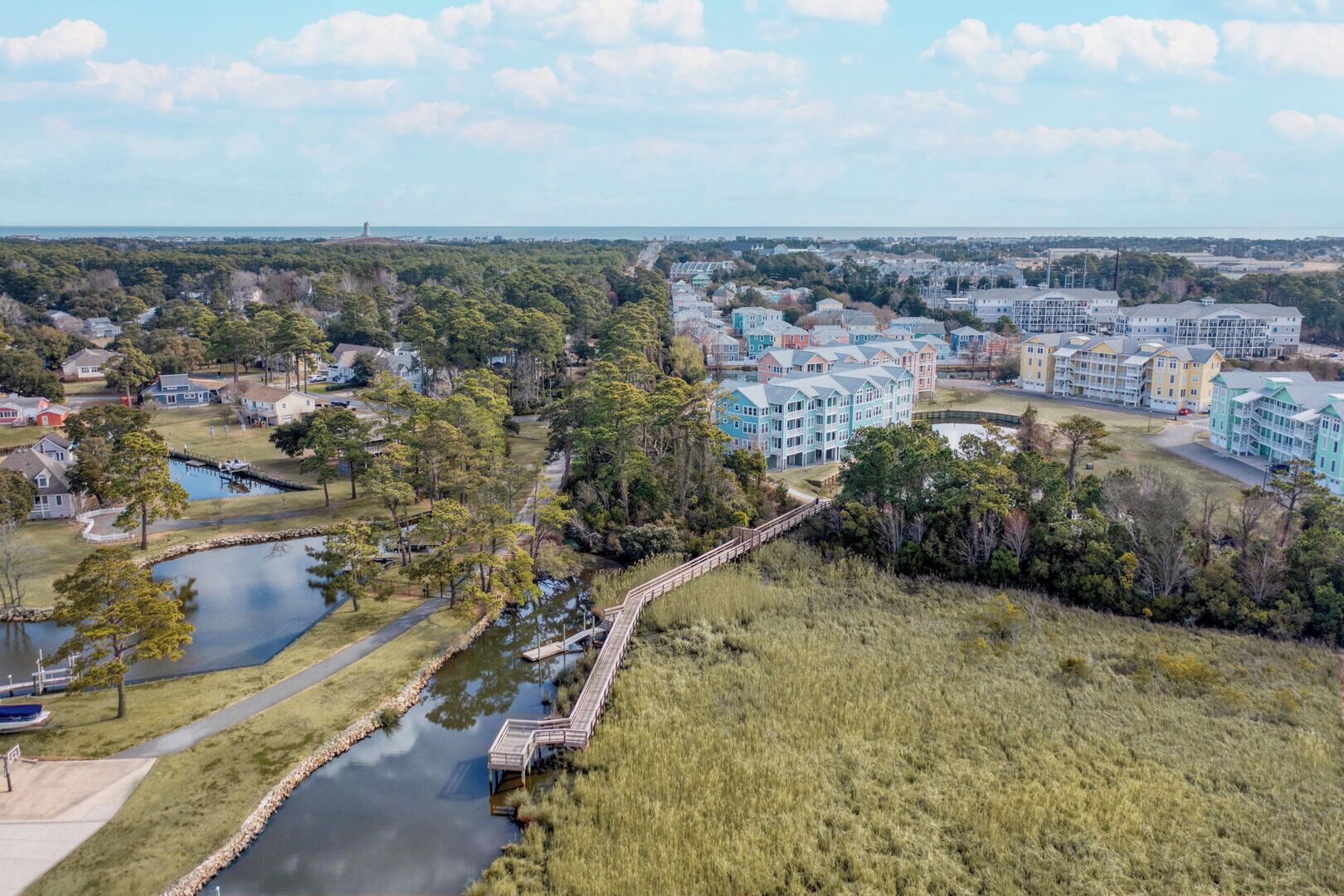 Zona residencial costera con casas unifamiliares y edificios de varios pisos, un pequeño puente sobre un canal de agua y una vista despejada del océano al fondo.