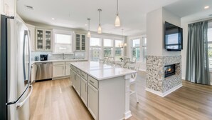 A modern kitchen with white cabinetry, a central island, pendant lighting, and a stone fireplace. - Turquoise Tide - Spacious End Unit, Sound Views & King En Suite (Kill Devil Hills)