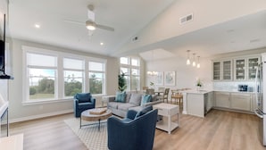 Un salon spacieux comprenant un canapé, des fauteuils et une table basse. Il y a également un coin cuisine avec des placards et un réfrigérateur. De grandes fenêtres laissent entrer la lumière naturelle et offrent une vue sur l’extérieur.