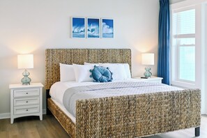 A neatly arranged bedroom with a woven headboard, a large bed with white linens, two bedside tables with lamps, and a window with blue curtains.