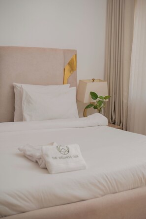 Un letto rifatto con lenzuola bianche, una testiera dorata, una lampada da comodino e un asciugamano con il logo 'The Wellness Club'.