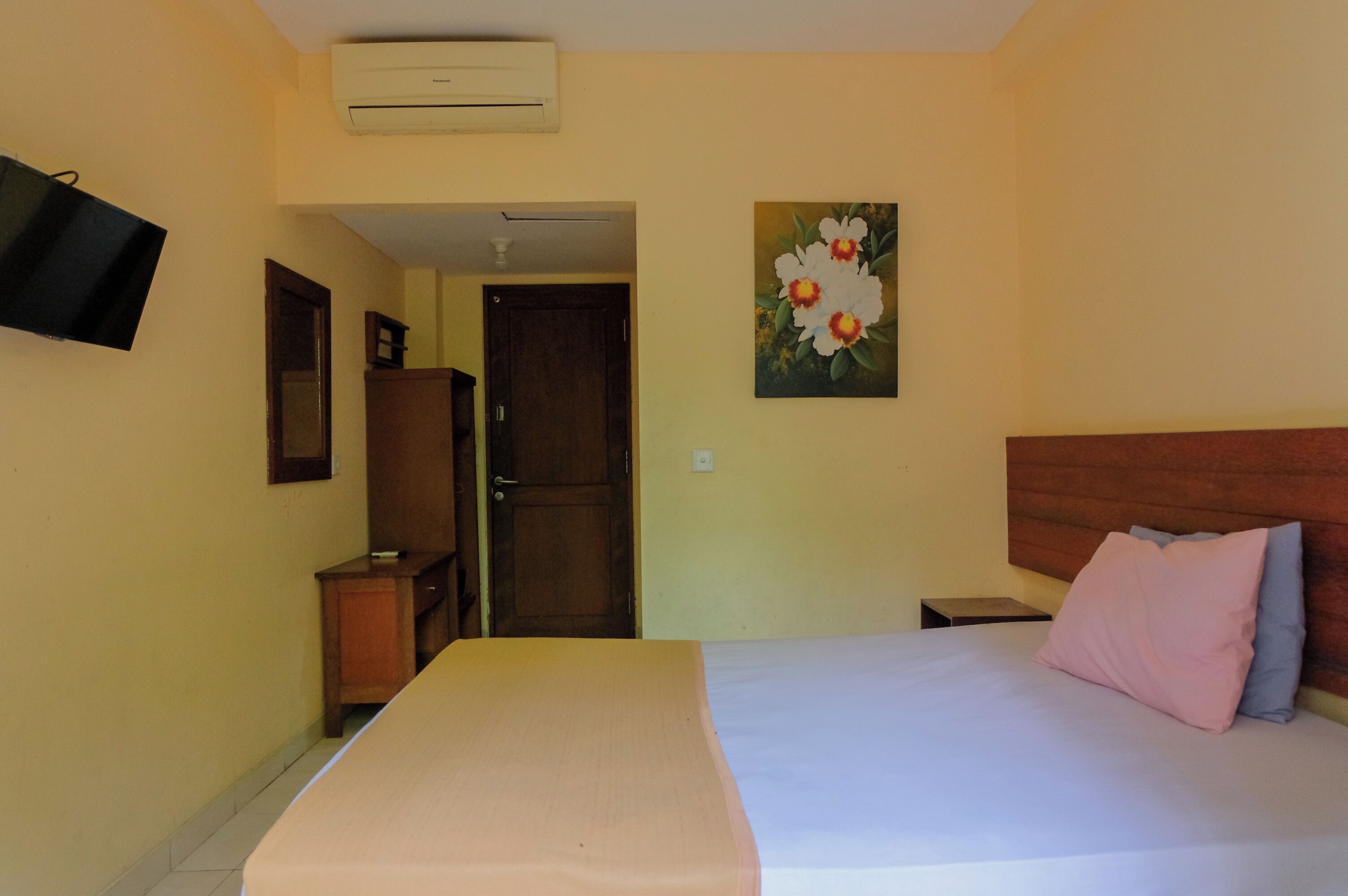 Una habitación de hotel con cama, televisor montado en la pared, una puerta y un cuadro de flores.