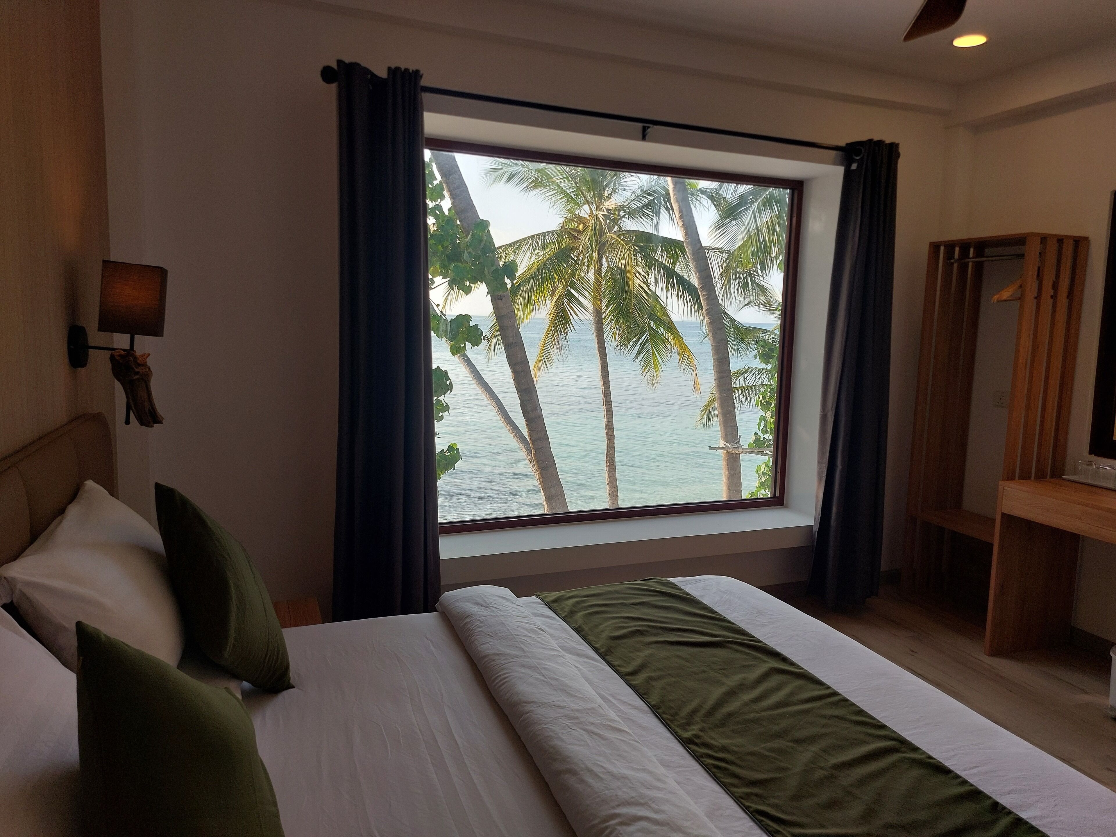 A hotel room with a large window, a bed with white bedding, green pillows, and a wooden nightstand.