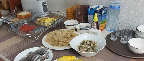 Ein Frühstücksbuffet mit verschiedenen Speisen, darunter Eier, Würstchen, Pfannkuchen und Früchte wie Bananen und Wassermelonenscheiben.