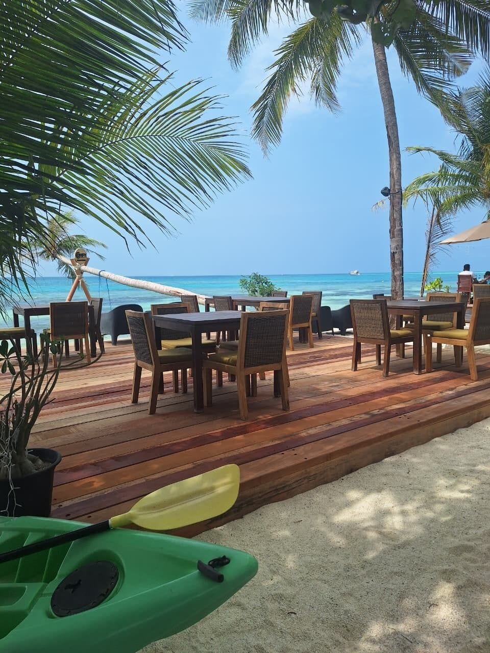 A wooden deck with tables and chairs, overlooking a beach with palm trees and the ocean.